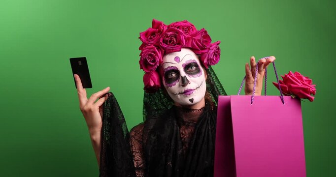 A girl wearing Calavera Catalina makeup, a flower crown, and lace mantilla holds a credit card in front of a green screen. She poses for Day of the Dead or Halloween.