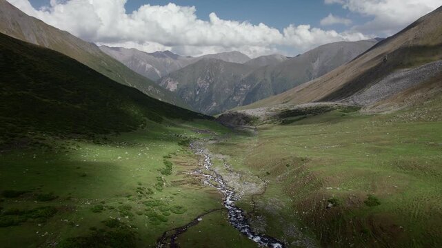 Aerial cinematic view of lush green valley surrounded by rugged rocky peaks in the remote Tien Shan mountain range, Kyrgyzstan.