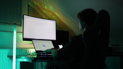 Man types on laptop in dark room with large monitor glowing and neon lights reflecting on wall concept of remote work online communication and digital lifestyle - Powered by Adobe