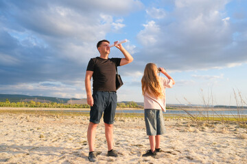 Dad and daughter are walking by the lake. The girl is looking through binoculars.