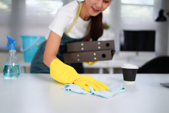 Young asian woman wearing yellow gloves and an apron, smiling while cleaning a white table with disinfectant spray and a blue cloth, promoting hygiene and cleanliness in her workspace