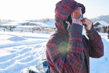 Caucasian young adult man adjusting ski goggles outdoors in snowy rural area, wearing hooded winter jacket, preparing for winter sports activity with snow covered landscape in background