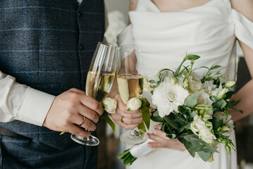 The bride holds a bouquet of white roses in one hand and a glass of sparkling wine in the other. The groom stands next to her and also holds a glass of sparkling wine