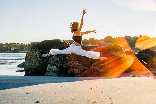 Graceful dancer performing splits leap over rocks on beach at sunset