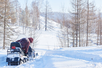 Man riding snowmobile through snowy forest landscape, navigating deep snow with trees in background, wearing winter clothing, facing away from camera