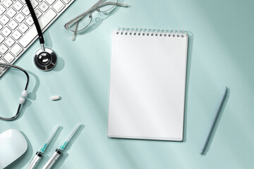 Doctor's desktop with blank notebook, glasses, stethoscope, syringe, pills, keyboard and mouse on light blue background with sunlight from the window.