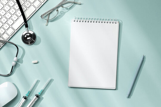 Doctor's desktop with blank notebook, glasses, stethoscope, syringe, pills, keyboard and mouse on light blue background with sunlight from the window.