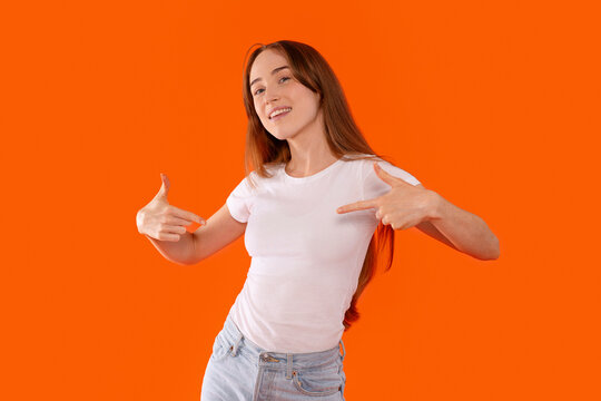 Young woman smiling and pointing at herself in front of a vibrant orange background during a playful photo session