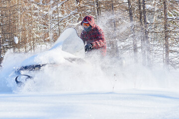 Man riding snowmobile through deep snow in winter forest, wearing protective goggles and hood, snow spraying around vehicle, trees in background