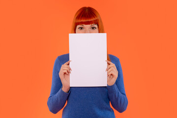 Young woman in blue sweater holds blank paper in front of her face against vibrant orange background during creative photo shoot
