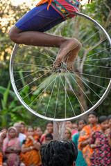 A young performer is balancing barefoot on a bicycle wheel, which is placed on an empty glass cup upon the head, while a fascinated crowd watches the daring act in an open-air village setting.