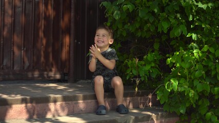 Child is happy and claps hands while sitting on the porch during a hot summer day in dark clothes
