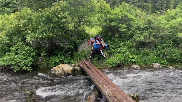 TTA tourist with a large backpacker and trekking poles crosses a stormy mountain river on logs. A trip to Kazakhstan. Kolsai lakes. Adventures in wild places