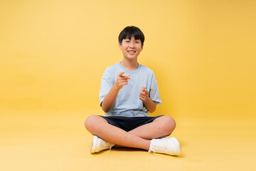 Fototapeta premium Smiling boy in casual clothes sits cross-legged on a yellow background pointing at the viewer