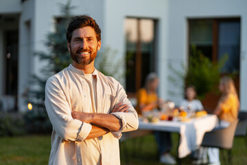 Smiling Hispanic man standing with arms crossed while family having dinner in the garden