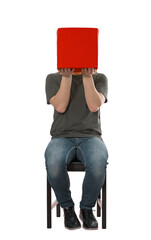Incognito young man seated on a chair holding a red box covering his face. Anonymous introvert person hiding identity behind a case, full length male portrait isolated on transparent background