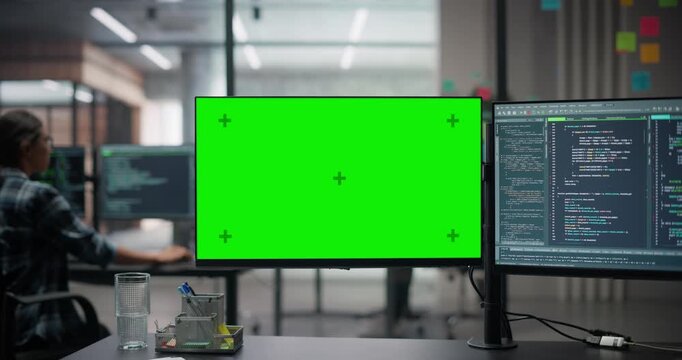 Office Desk with Two Computer Monitors in a Technology Office with a Female Working in the Background. Green Screen Mock Up Template on One Screen, Software Running on Second Display