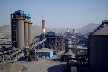 Obraz premium Large industrial mining complex with multiple structures, silos, conveyor belts, and smokestacks emitting smoke, set against mountainous background, clear sky, showcasing heavy machinery