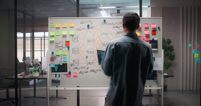 Young Man Walking Up to an Office Whiteboard with a Laptop Computer. Project Manager Reviewing Information, Looking at the Technical Data and Graphs. Footage from Behind