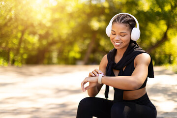 Cheerful sporty African American woman with headphones checking smartwatch while smiling outdoors. Fitness tracking, digital lifestyle, health and motivation concept
