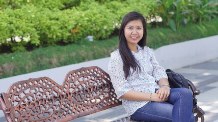 Young Asian woman sitting on a decorative bench in the park, wearing a casual floral blouse and jeans, enjoying a relaxing moment outdoors with natural greenery background