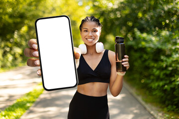 Happy young sporty African American woman showing smartphone with blank screen outdoors, holding water bottle. Fitness app, wellness, technology and active lifestyle, mockup