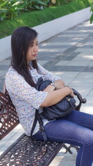 Young Asian woman sitting on a decorative bench in the park, wearing a casual floral blouse and jeans, enjoying a relaxing moment outdoors with natural greenery background