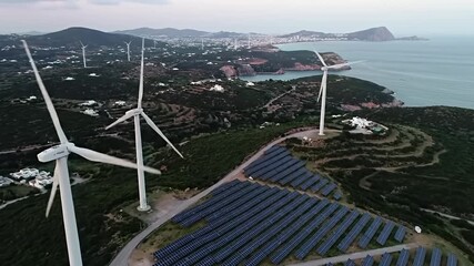 Aerial view of wind turbines and solar panels on a hillside overlooking a coastal town - Powered by Adobe