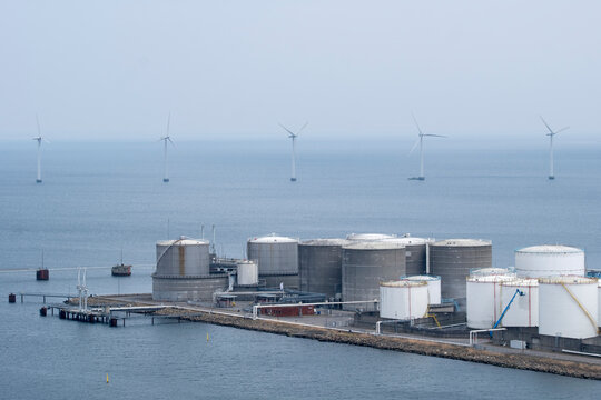 Coastal storage tanks in Copenhagen harbor with offshore wind turbines in background, juxtaposing fossil based industry with renewable energy presence in maritime landscape