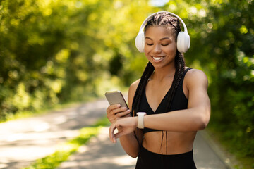 Smiling sporty African American woman with headphones checking smartwatch outdoors. Fitness tracking, technology, lifestyle and wellness during active routine