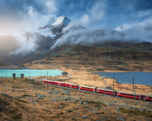 Aerial view of red Bernina Express train, curving railroad, turquoise lake in the Swiss Alps, rocky hills and dramatic cloudy sky at sunset in autumn. Alpine mountains. Top view of train in fall