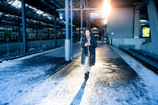Confident middle aged woman in winter coat walking on snowy train platform with phone, urban commuting, cold season lifestyle in bright daylight.