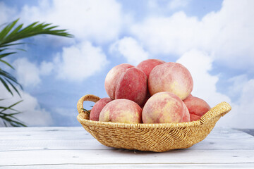Fresh Ripe Peaches in Wicker Basket on White Wood Table with Blue Sky Background