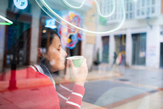 Woman with dark hair enjoying hot drink by cafe window, eyes closed in peaceful moment. Urban reflections, cozy lifestyle and relaxation in modern city environment