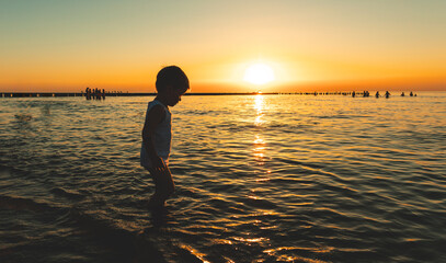 Little boy plays in the shallow water on a sandy beach during sunset on a warm summer evening