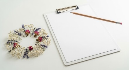 A blank clipboard with paper and a pencil lies on a white surface next to a circular wreath of dried pink rosebuds white blooms and purple lavender sprigs