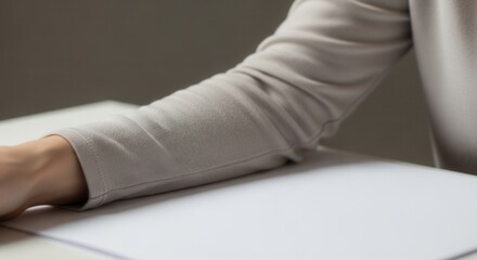 A persons arm in a light long-sleeved top rests on a white table next to a blank sheet of paper