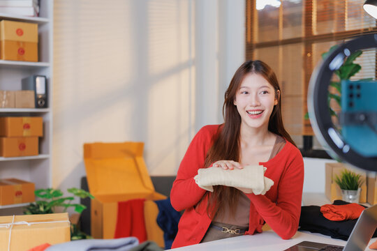 Young asian female entrepreneur showing clothes while recording a video for selling online from her home office, surrounded by cardboard boxes and using a ring light