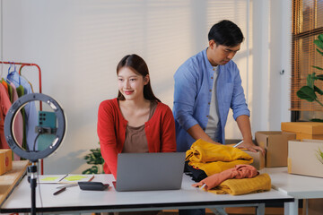 Two young Asian entrepreneurs collaborating in a home office, preparing clothing for shipping while engaging in live streaming to promote their online sales and connect with customers