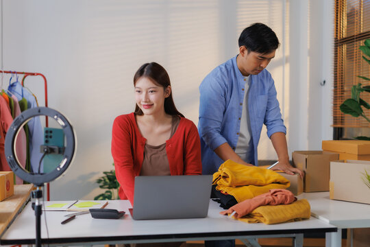 Two young Asian entrepreneurs collaborating in a home office, preparing clothing for shipping while engaging in live streaming to promote their online sales and connect with customers