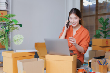 Young Asian woman entrepreneur talking on the phone while preparing packages for delivery in a home...