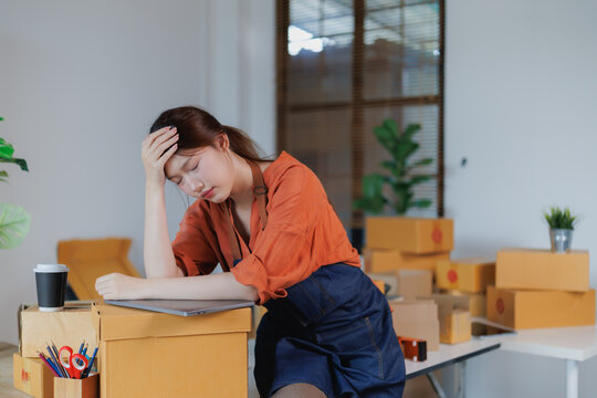 Young Asian woman entrepreneur feeling tired and stressed while working on a laptop in her shop, surrounded by cardboard boxes and struggling with online business results