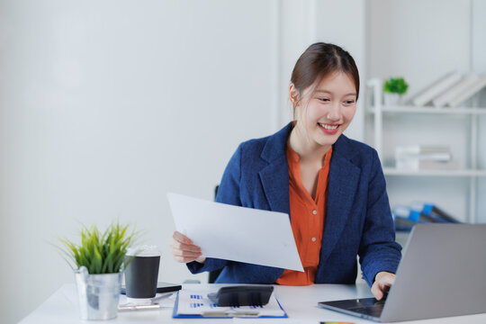 Smiling asian businesswoman working with laptop and analyzing financial document, sitting at her workplace in modern office with charts and graphs, plant and calculator on desk