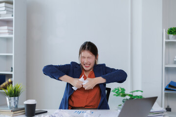 Asian businesswoman ripping paper in anger while sitting at office desk with laptop and crumpled papers, expressing frustration and stress in workplace