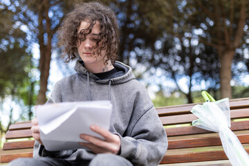Young man reading documents on park bench with folded umbrella