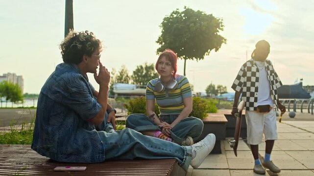 Long shot of tattooed red-haired gen Z girl sitting on bench next to male Asian peer while gossiping about African American skateboarder passing them by on city street