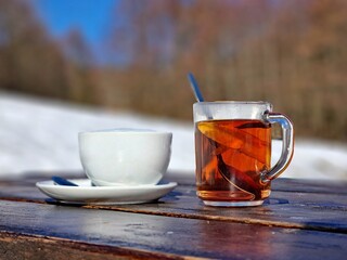 Winter outdoor scene with a glass of tea and a cappuccino cup on a wooden table, snow-covered background, blue sky, perfect cozy coffee moment.