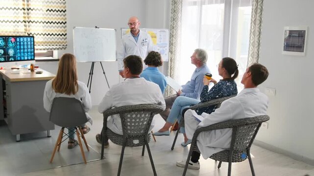 Adult bald professor in white coat explains new drugs during hospital seminar. Lecturer points at board with medical notes while healthcare staff attend professional training session.