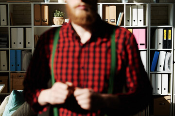 Male hipster in office. Bearded man in checkered shirt and suspenders out of focus standing against office shelves with file holders