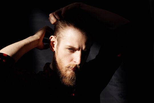 Portrait of young bearded man standing in dark room, smoothing his hair and looking at camera piercingly, his face illuminated by bright sun